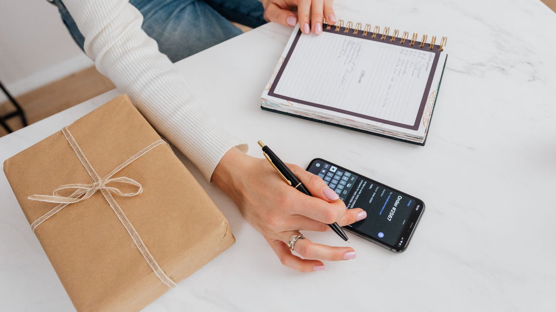 A hand with a pen taps a phone beside an open notebook and a gift wrapped in brown paper and twine
