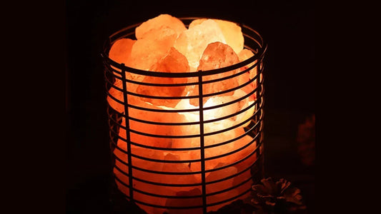 A cylindrical black wire basket lamp filled with rough chunks of pink rock salt, lit amber in a dark room