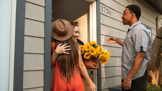 A visitor in a brown hat hugs a host at a front door while a guest holds a pot of sunflowers
