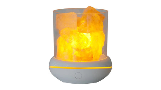 A small USB salt lamp with pale pink salt chunks in a clear glass cylinder, glowing amber on a light grey base with a single button