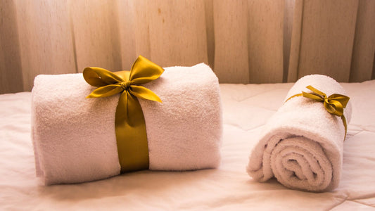 Two rolled white towels tied with gold satin ribbons on a made bed in front of a cream curtain