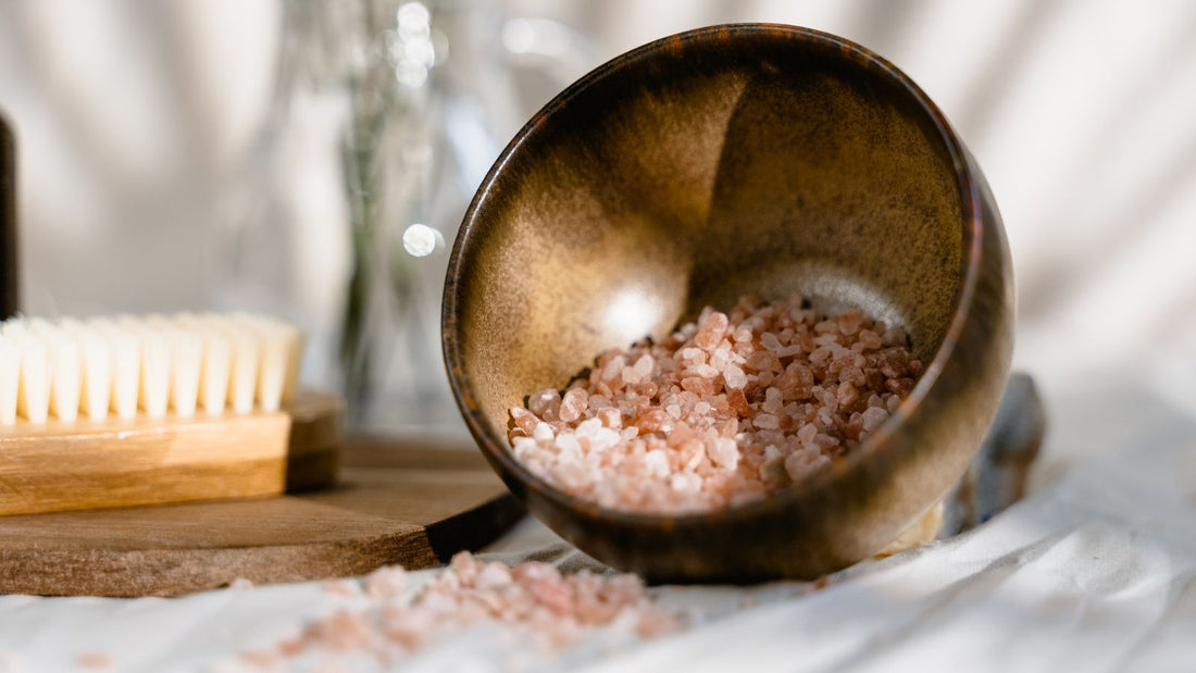 Pink Himalayan salt spilling from a dark ceramic bowl beside a wooden brush in soft daylight