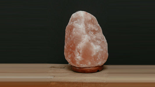 A large natural-shape pink salt lamp on a round wooden base, unlit, on a wooden bench against a dark wall