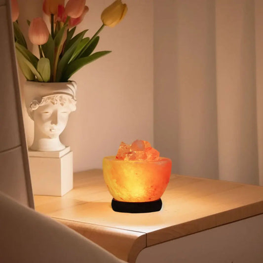 A bowl-style Himalayan salt lamp glowing on a wooden side table beside a plant and a ceramic bust