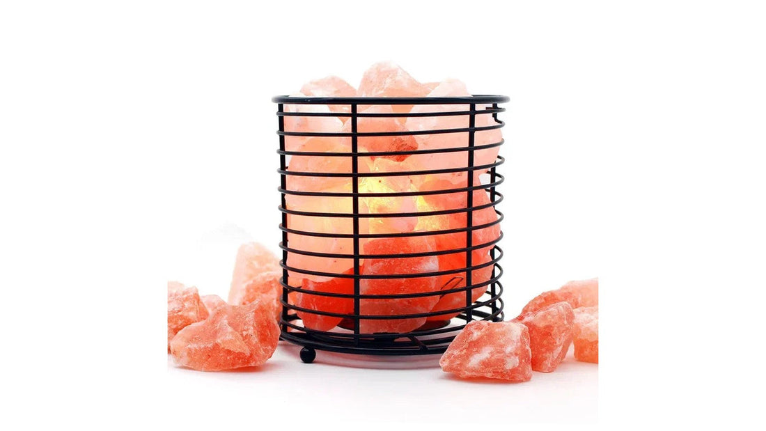 A wire-cage Himalayan salt lamp lit on a white surface with loose pink salt chunks unpacked around it